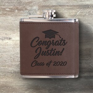 May include: A brown leather flask with a silver cap. The flask is engraved with the words "Congrats Justin!" and "Class of 2020" along with a graduation cap graphic. A graduation gift.