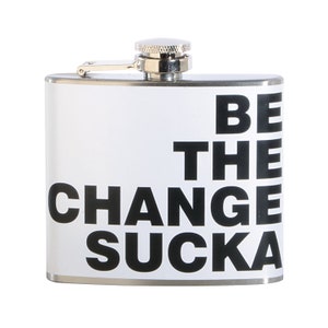 Op de afbeelding: Een zilveren roestvrijstalen hipflask met een witte achtergrond en zwarte tekst die "BE THE CHANGE SUCKA" luidt.