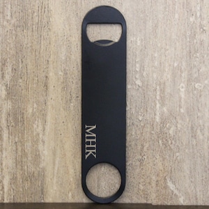 May include: Black bottle opener with a rectangular body, a circular opening at the bottom, and a bottle cap opener at the top. The initials "MHK" are engraved vertically on the body.