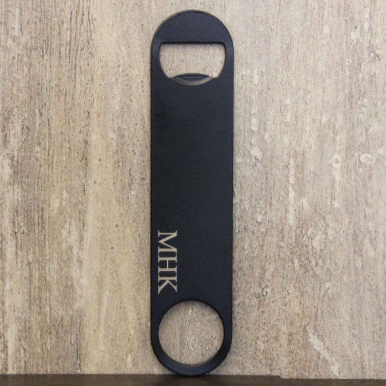 May include: Black bottle opener with a rectangular body, a circular opening at the bottom, and a bottle cap opener at the top. The initials "MHK" are engraved vertically on the body.