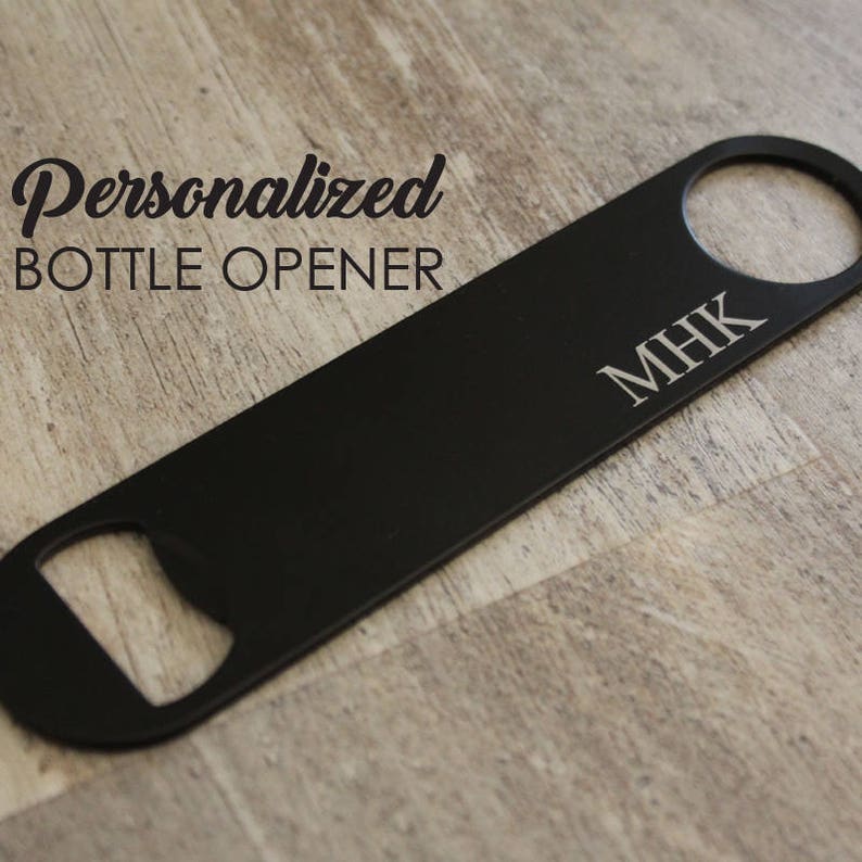 May include: A black, personalized bottle opener with the initials "MHK" engraved on the surface. The text "Personalized BOTTLE OPENER" is printed in a script font. The opener has a circular hole at one end and a bottle cap opener at the other.