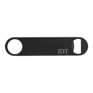 May include: A black bottle opener with a circular hole on one end and a bottle cap opener on the other. The initials "JDT" are printed in the center of the opener.