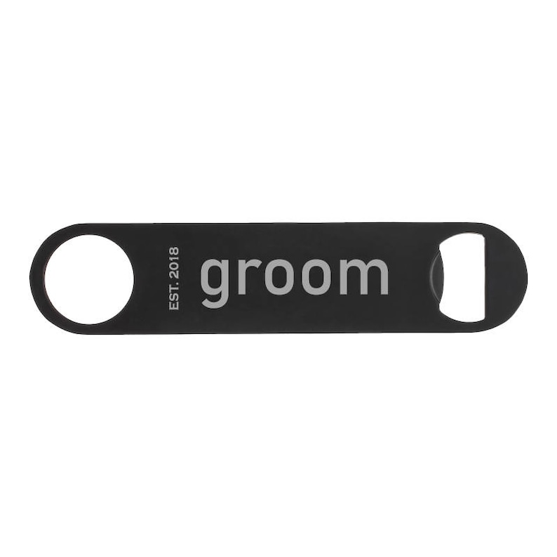 May include: A black bottle opener with a circular hole on one end and a bottle cap opener on the other. The word "groom" is printed in light gray, along with "EST. 2018".
