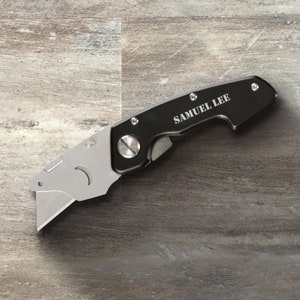 May include: A black utility knife with a silver blade and a personalized inscription that reads "SAMUEL LEE".