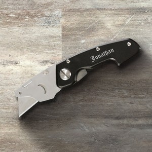 May include: A black utility knife with a silver blade and a personalized inscription that reads "Jonathan".