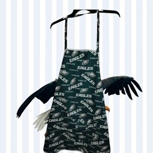 May include: A dark green apron with the word "EAGLES" and a feather design repeated across the fabric. The apron is hanging on a black hanger. The background is a light blue and white striped pattern.