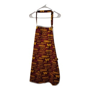 May include: A burgundy apron with a repeating pattern of the Washington Commanders logo and text. The apron is hanging on a white hanger against a white background. The apron has a neck strap and a waist tie.