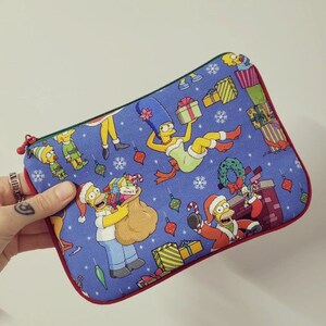 Puede incluir: Una bolsa rectangular azul con cremallera, con un diseño navideño con personajes de Los Simpson. El diseño incluye a Homer, Marge y otros personajes, junto con copos de nieve, regalos y adornos. La bolsa tiene un ribete rojo y una cremallera verde.