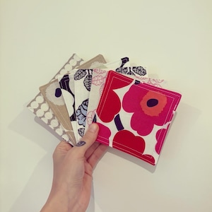 May include: A hand displays a selection of square fabric coasters with diverse floral and geometric designs. The coasters showcase colours including red, pink, white, black, and beige. The coasters are made of fabric and have a stitched edge.