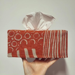 Puede incluir: Una funda para caja de pañuelos con estampado geométrico rojo y blanco. La funda está hecha de tela y tiene una caja de pañuelos blanca en su interior.