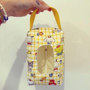 Könnte beinhalten: Eine gelbe Taschentuchbox-Hülle mit einem sich wiederholenden Muster von Cartoon-Charakteren und einem gelben Karomuster. Die rechteckige Hülle hat eine große Öffnung und einen gelben Griff.