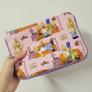 Puede incluir: Una bolsa rosa con cremallera y ribete morado con un patrón repetido de los personajes de Los Simpson. La bolsa tiene el texto "The Simpsons" impreso.