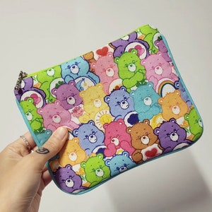 Może przedstawiać: Turkusowa kosmetyczka zapinana na zamek błyskawiczny z kolorowym nadrukiem Care Bears. Kosmetyczka jest wypełniona Care Bears w różnych kolorach, w tym różowym, niebieskim, żółtym, zielonym i fioletowym. Każdy Care Bear ma inny wyraz twarzy i trzyma serce.