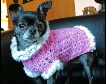 May include: A small black chihuahua wearing a pink crocheted sweater with a white fur trim. The dog is sitting on a black couch.