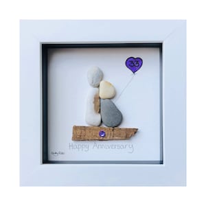 May include: A framed pebble art piece with a white frame. The art depicts two figures made of pebbles, a purple heart balloon with the number 33, and the words "Happy Anniversary". A small purple gem is on the driftwood base.