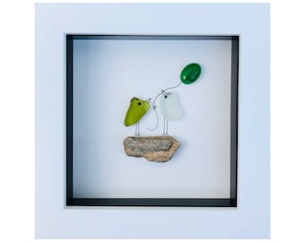 May include: A framed artwork features two glass birds on a piece of driftwood. One bird is lime green, the other is white, and they are connected by a wire to a green glass balloon. The frame is white.