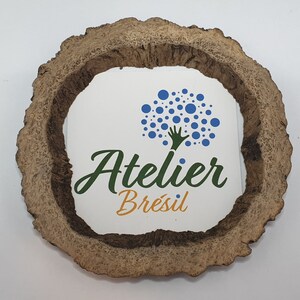 Può includere: Un sottobicchiere rotondo in legno con un adesivo bianco che presenta un logo blu e verde. Il logo rappresenta un albero stilizzato con una mano che si estende verso l'alto. Il testo "Atelier Brésil" è scritto sotto l'albero.