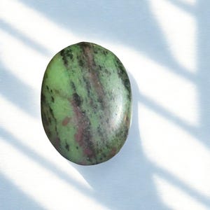 Może przedstawiać: Owalny kamień szlachetny z nakrapianym wzorem w odcieniach zieleni, czerni i brązu. Wypolerowany kamień umieszczono na białym tle z cieniami, podkreślając jego naturalne kolory i faktury. Gładka powierzchnia kamienia szlachetnego odbija światło.
