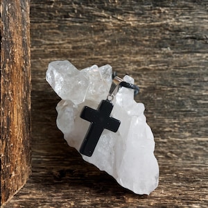 May include: A black cross pendant with a silver-colored bail hangs on a black cord. The cross rests on a cluster of clear quartz crystals. The background is a rustic wooden surface.