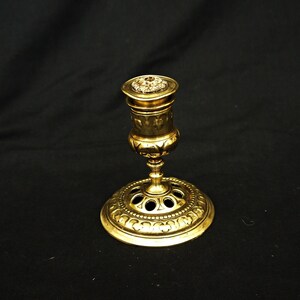 May include: A gold metal candlestick with a round base and a decorative design. The candlestick has a small, round top with a floral design.