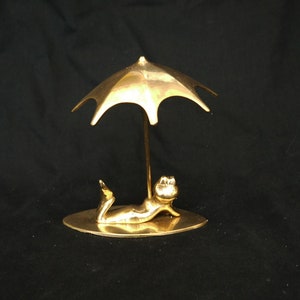 May include: A gold-colored metal figurine of a person lying down under a small umbrella. The figurine is on a small, oval-shaped base.