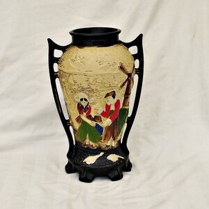 May include: A ceramic vase with black handles and a cream-coloured body. The vase features a painted scene of two people in traditional clothing standing in front of a windmill.
