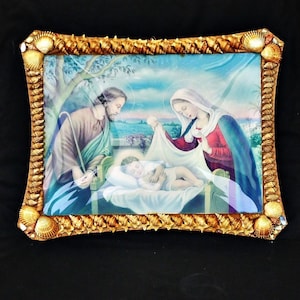 Può includere: Un dipinto incorniciato che rappresenta la scena della Natività con Maria, Giuseppe e il bambino Gesù. La cornice è fatta di conchiglie e vernice dorata.