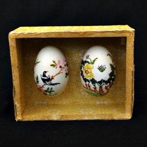 Puede incluir: Dos huevos blancos decorativos con diseños florales y de aves pintados, presentados en una pequeña caja amarilla. Un huevo presenta un pájaro negro en una rama con flores rosas, el otro tiene flores amarillas y follaje verde.