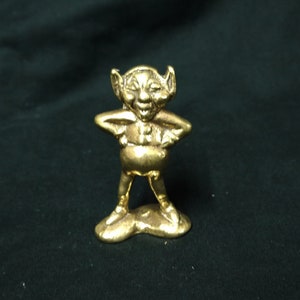 May include: A small, gold-coloured figurine of a smiling gnome with outstretched arms. The gnome is standing on a small, gold-coloured base.