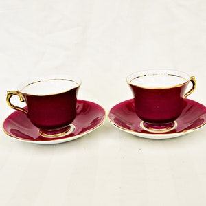 May include: Two burgundy teacups and saucers with gold trim. The teacups have a classic shape with a handle. The saucers are round and match the color of the teacups. The interior of the teacups is white.
