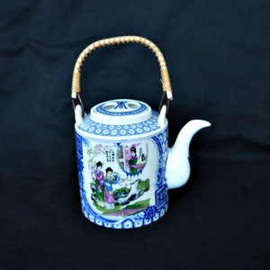 Op de afbeelding: Een blauwe en witte porseleinen theepot met een bamboe handvat. De theepot heeft een bloemenmotief en een scène met twee vrouwen in traditionele kleding.
