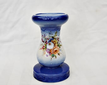 May include: A blue and white ceramic candle holder with a floral design. The holder has a wide base and a narrow neck.