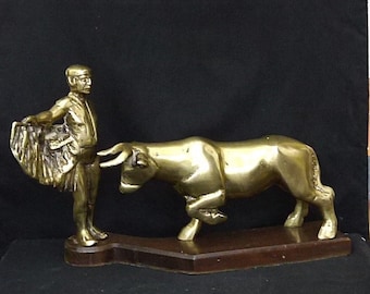 May include: A gold-toned metal sculpture of a bullfighter and a bull on a wooden base. The bullfighter is holding a cape and the bull is charging.