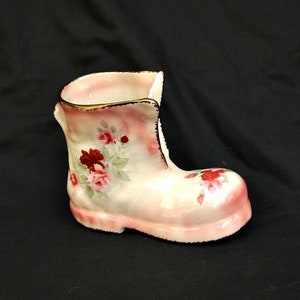 Pode incluir: Um vaso de cerâmica branca em forma de bota com um esmalte rosa e um design floral. O vaso é decorado com rosas vermelhas e rosas e folhas verdes.