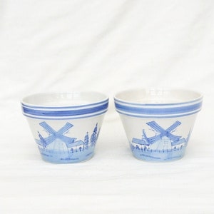 May include: Two white ceramic flower pots with blue and white windmill designs. The pots have a blue stripe around the top.