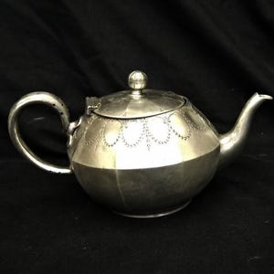 Puede incluir: Una tetera de plata con un diseño decorativo en el cuerpo y la tapa. La tetera tiene un cuerpo redondo y un pico curvo.
