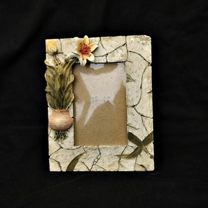 Op de afbeelding: Rechthoekige fotolijst met een gebarsten, gebroken witte afwerking. Het frame is versierd met een kleine pot met planten en een bloem. Het ontwerp van het frame bevat groene bladeren en een neutraal gekleurd interieur.