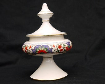 May include: A white ceramic candy dish with a gold trim and a floral design. The dish has a lid and a pedestal base.
