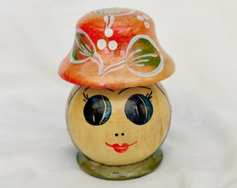 May include: A wooden doll with a painted face, large eyes, and a red, white, and green hat. The doll is a traditional Russian nesting doll.
