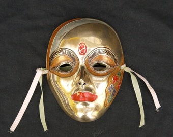 May include: A gold-coloured masquerade mask with pink and white ribbons. The mask has a painted design of flowers and a red dot on the forehead.