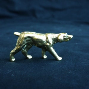 May include: A gold-colored metal figurine of a dog walking, with its head turned to the right. The figurine is on a dark blue background.
