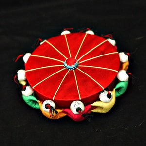 May include: A red circular pincushion with a white starburst design and a blue center. The pincushion is surrounded by a ring of small, colorful figures with black eyes.