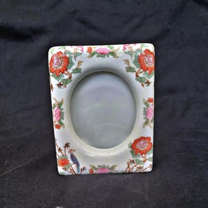 Puede incluir: Un marco de fotos rectangular de cerámica blanquecina con una abertura ovalada. El marco está decorado con motivos florales rojos y rosas y un pájaro azul. Es un artículo decorativo.