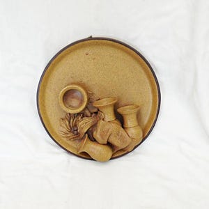 May include: A circular, tan ceramic wall plaque with a dark brown border. The relief design showcases small vases and a central circular aperture. The piece has a rustic, earthy aesthetic, suitable for home decor.
