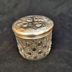 May include: An antique glass jar with a silver lid. The lid is decorated with a relief of cherubic faces. The jar's body has a cut-glass pattern. The jar is likely for storing small items.