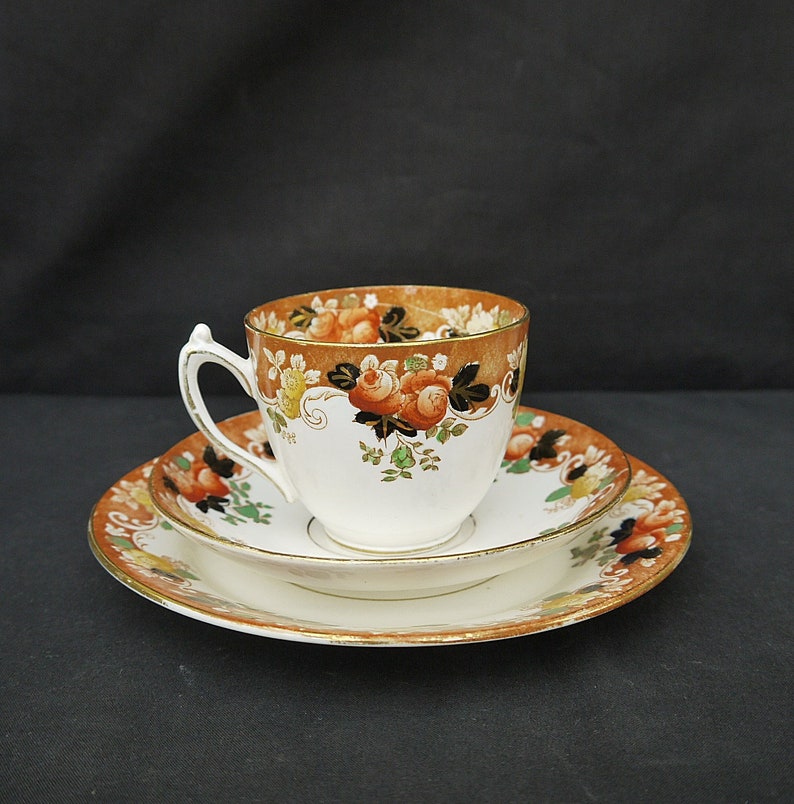 May include: A vintage teacup and saucer set with a floral pattern in shades of orange, brown, green, and black. The cup and saucer have a gold trim.