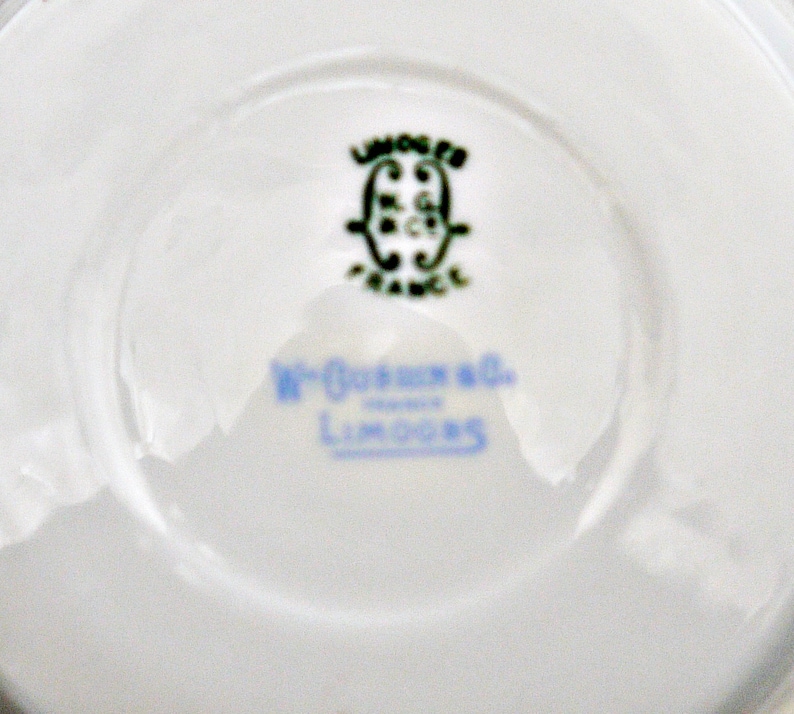 Puede incluir: Primer plano de la parte inferior de un platillo de porcelana blanca con un logotipo verde y negro que dice "Limoges France" y una inscripci&oacute;n azul que dice "W. Goerz & Co. Limoges".