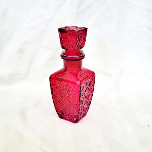 Può includere: Un decanter in vetro rosso rubino con base quadrata e tappo decorativo. Il decanter presenta un motivo geometrico ed è alto circa 15 cm.