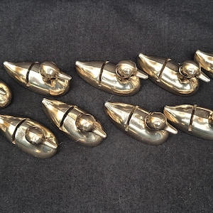 May include: A collection of ten polished brass duck-shaped knife rests arranged in two rows. Each small metallic figure features a smooth, reflective surface and a distinct vertical slit across the back, designed to hold a knife blade securely.