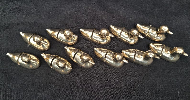 May include: A collection of ten polished brass duck-shaped knife rests arranged in two rows. Each small metallic figure features a smooth, reflective surface and a distinct vertical slit across the back, designed to hold a knife blade securely.